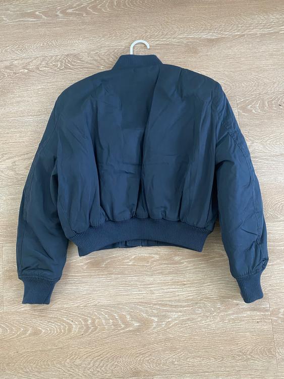 Black bomber jacket hanging on a hanger over a light wooden floor, viewed from behind.