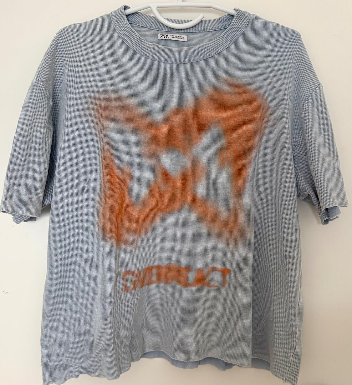 Light blue T-shirt with orange spray paint 'X' design and "OVERREACT" text at the bottom.