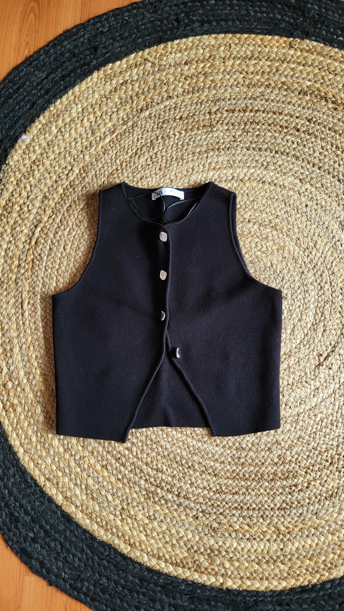 Black gilet with buttons on a round jute rug in beige and black tones, placed on a wooden floor.
