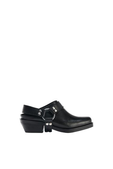 Black low-heeled shoe with straps and metallic details on a white background.