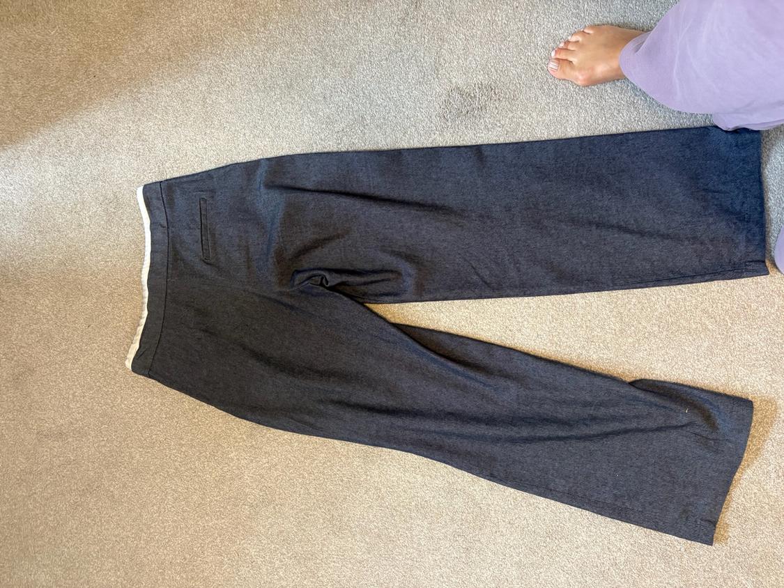 Dark grey wide-leg trousers on a light-coloured carpet. In the top right corner, a bare foot and part of a lilac garment can be seen.