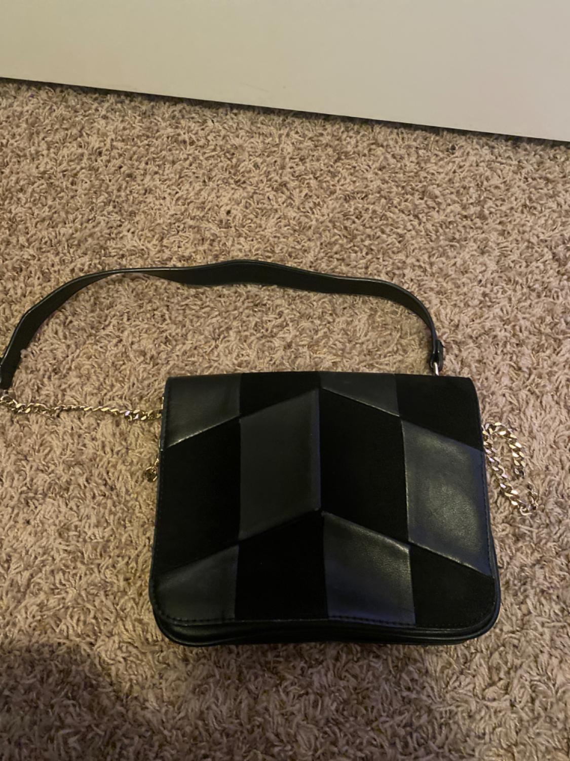 Black handbag with a geometric design on the beige carpeted floor. It has a gold chain strap and a leather strap.