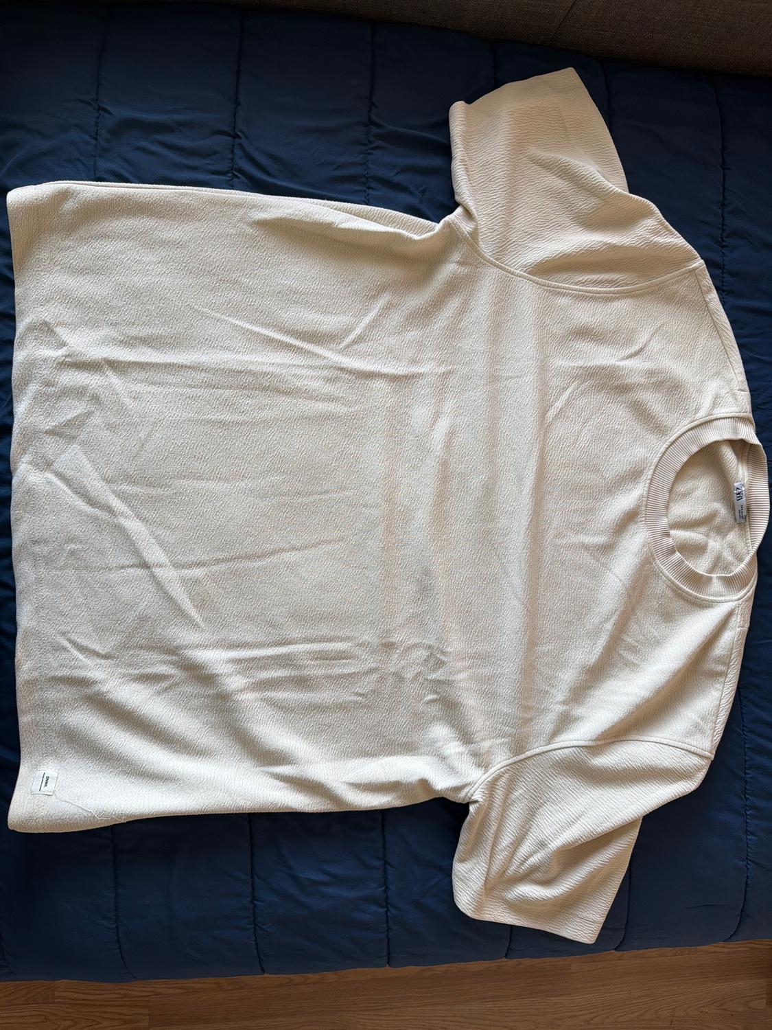 White short-sleeved T-shirt on a blue bedspread. The fabric has a light texture and is laid out flat.