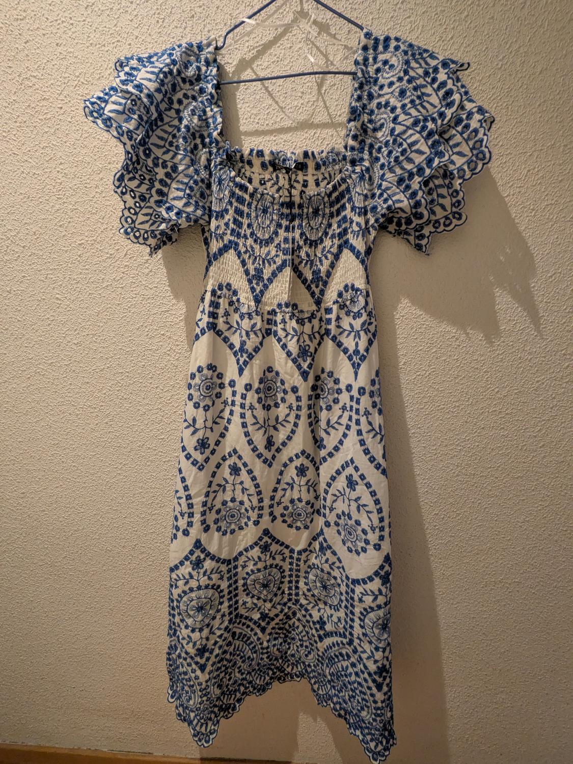 White dress with blue embroidery, frilled sleeves and a square neckline, hanging on a blue hanger against a textured wall.