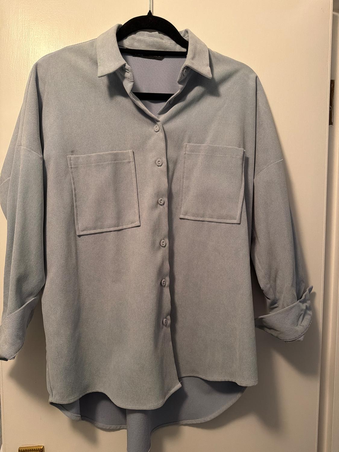 Light grey, long-sleeved shirt with two front pockets and buttons, hanging on a black hanger over a white door.