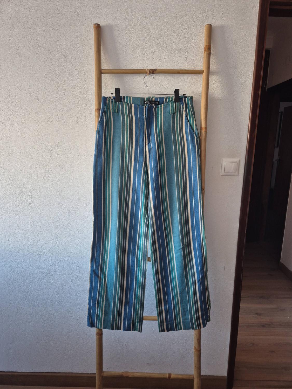 Wide blue, green and white vertical striped trousers, hanging on a wooden ladder leaning against a white wall.