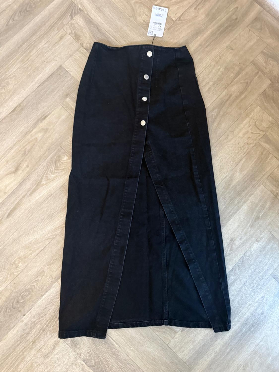 Black denim maxi skirt with front buttons and a central slit, laid out on a light wood floor with a herringbone pattern.