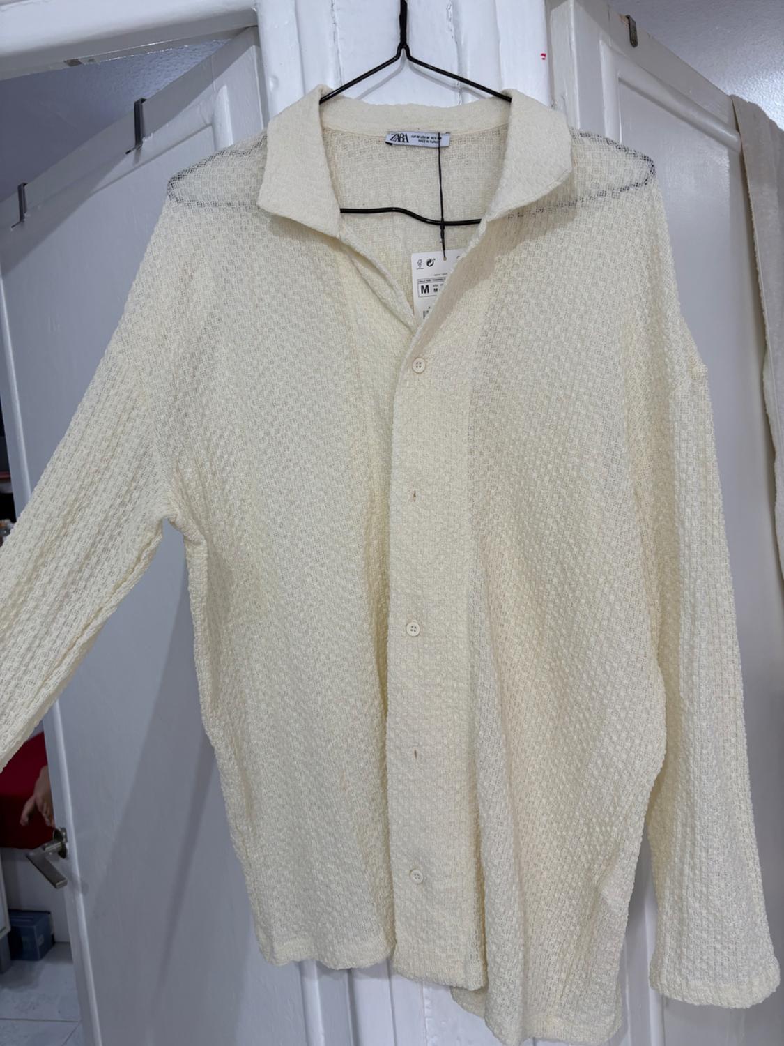 Beige textured shirt hanging on a black hanger, with a visible size M label. White doors in the background.