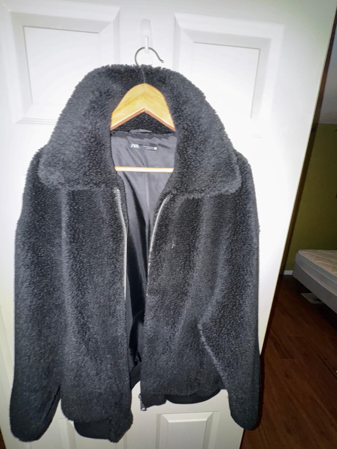 Black fluffy textured jacket hanging on a wooden hanger over a white door. Visible label: ZARA.