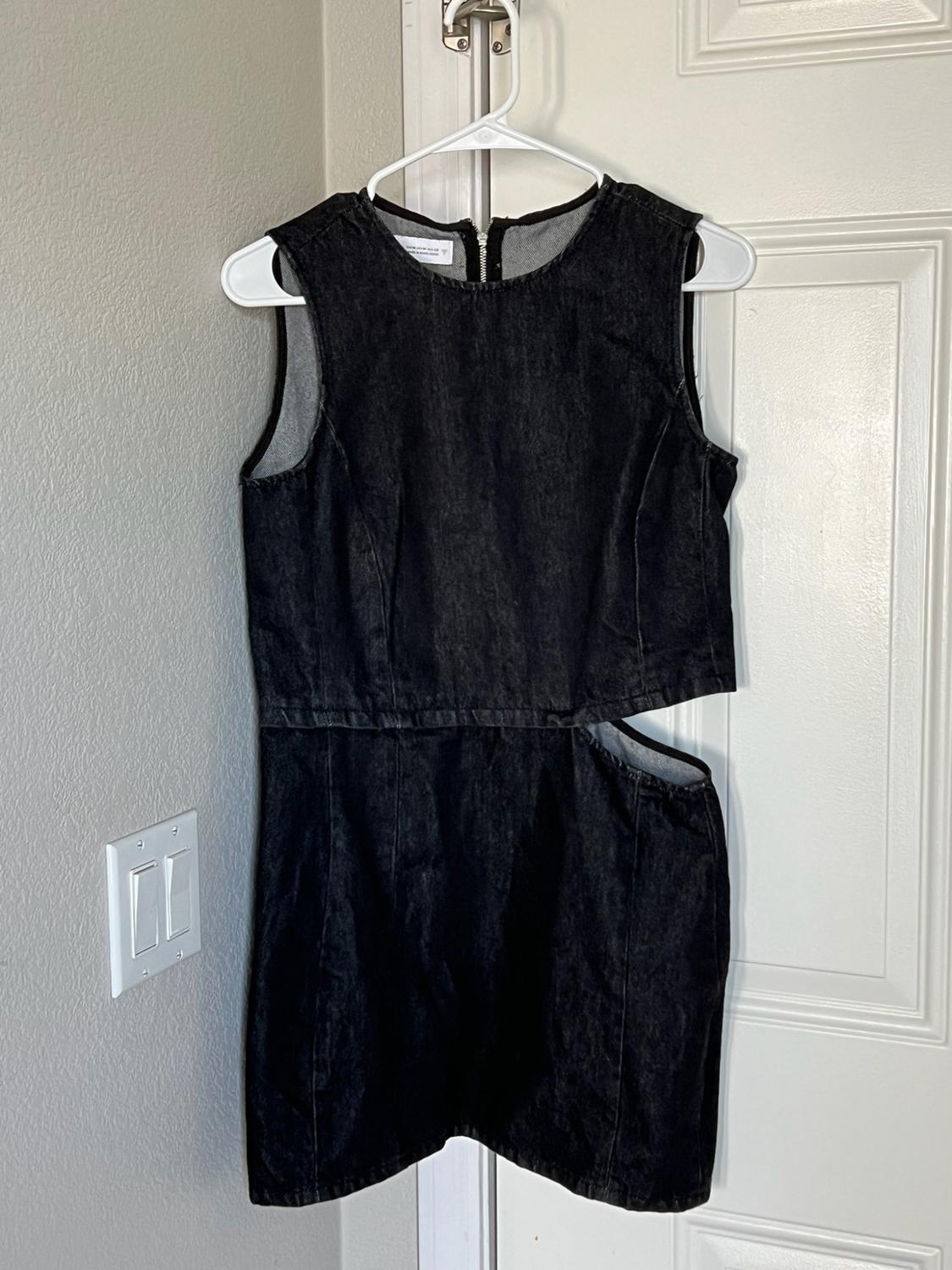Black sleeveless denim dress with a side slit, hanging on a hanger on a white door next to a wall with a light switch.