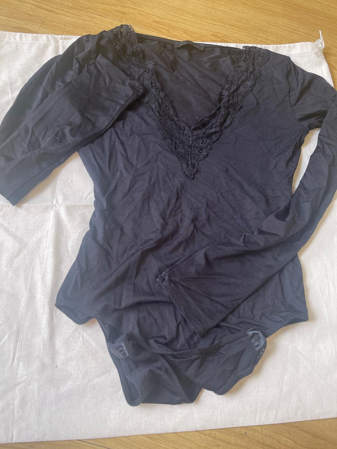 Black long-sleeved bodysuit with a lace V-neck, on white fabric and a light wood background.