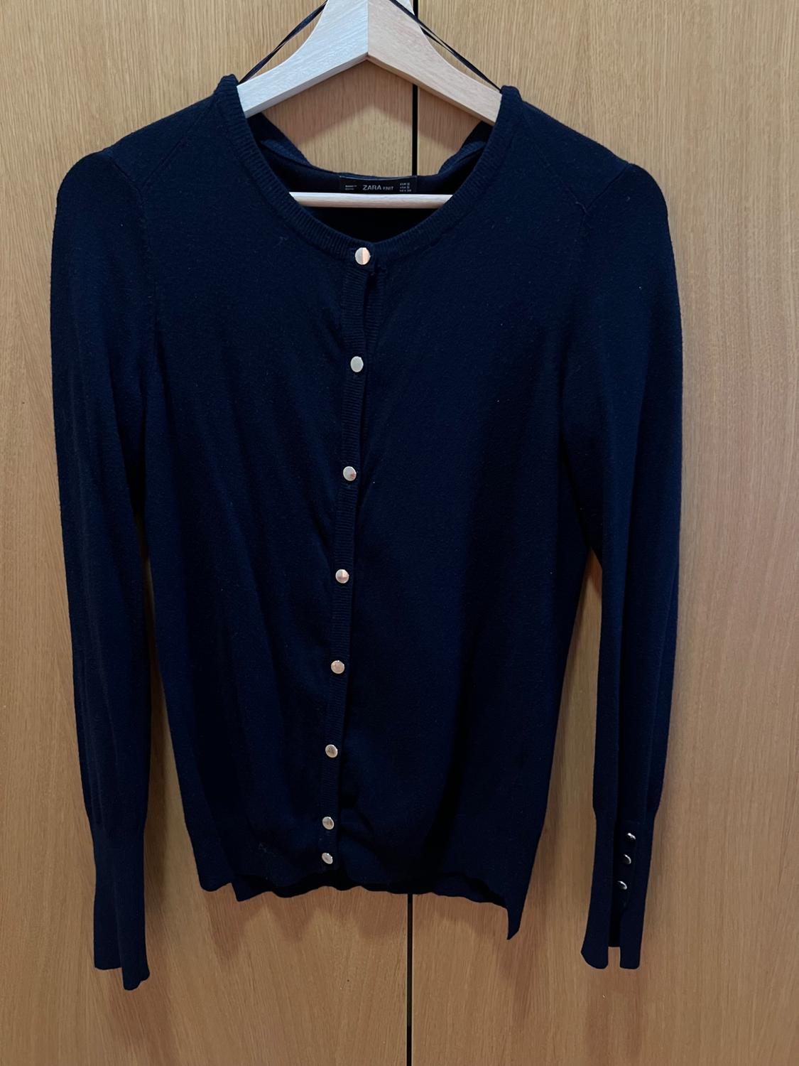 Navy blue knitted cardigan with metallic buttons, hanging on a hanger against a wooden background. Label visible: ZARA KNIT.