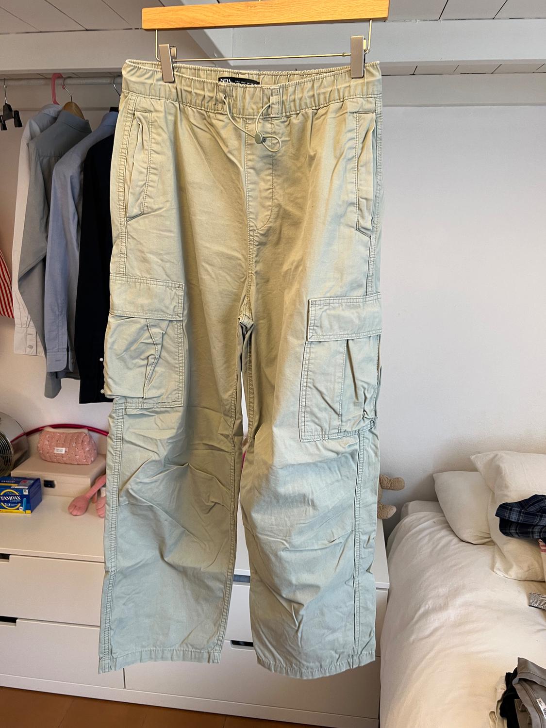 Beige cargo trousers hanging on a hanger in a bedroom. Background with white bed and clothes hanging in an open wardrobe.