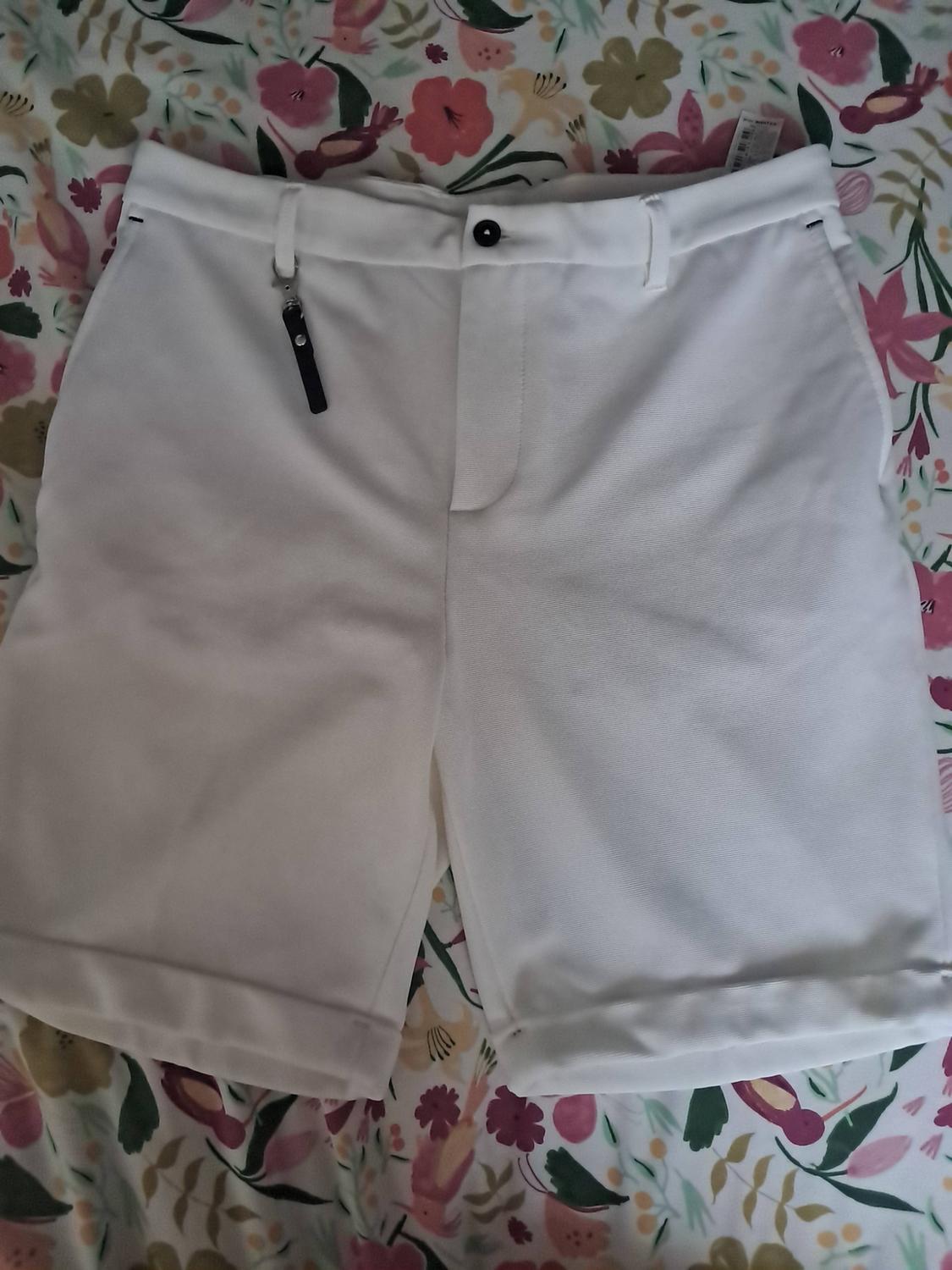 White shorts with a hem, dark button and black strap on the belt loop, set against a fabric background with a floral and bird print.