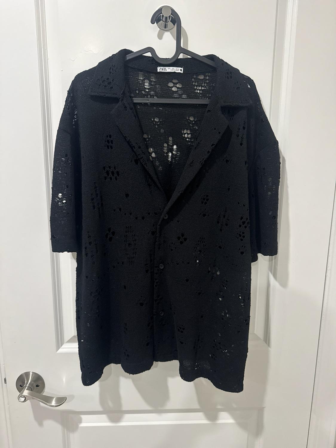 Black short-sleeved shirt with perforated fabric and buttons, hanging on a hanger over a white door. Visible text: ZARA.