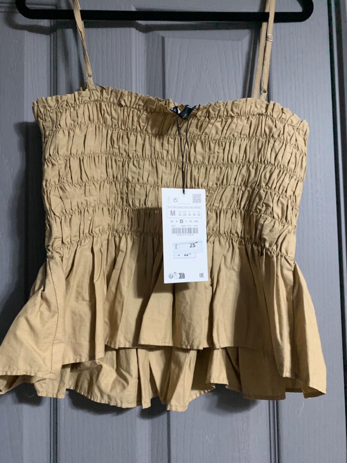 Beige top with spaghetti straps and ruched design, hanging on a black hanger. Visible tag with a price of £25.