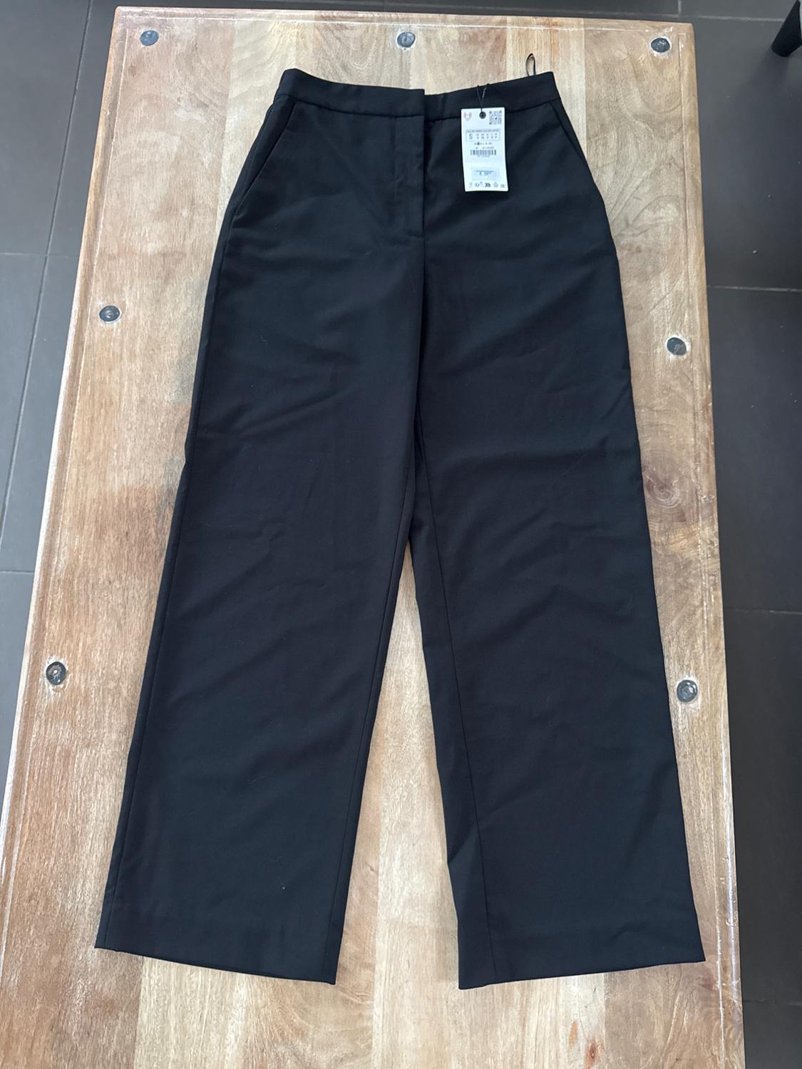 Black wide-leg trousers on a wooden table, with a visible label on the waistband.