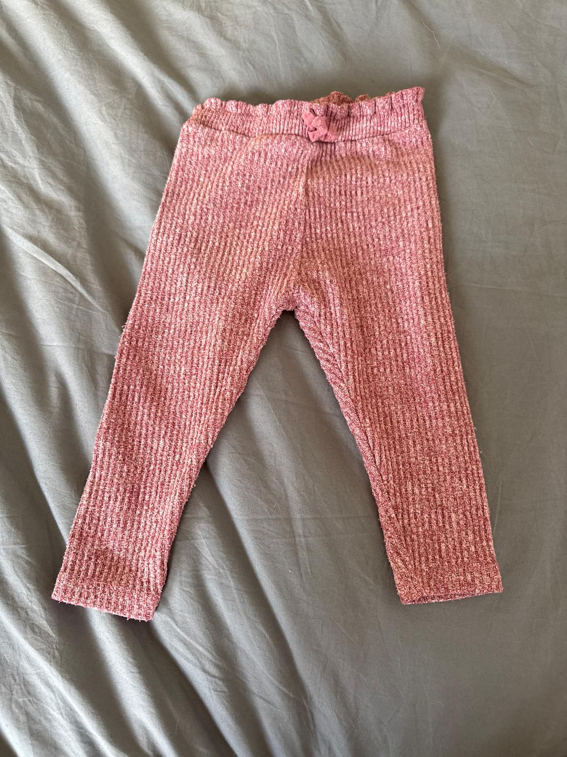 Pink knitted baby trousers on a grey surface. Ribbed design with elasticated waist and decorative bow.