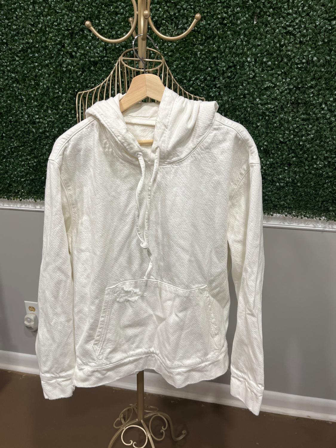 White hoodie with a front pocket hanging on a gold clothes rack, green wall background with leaf texture.