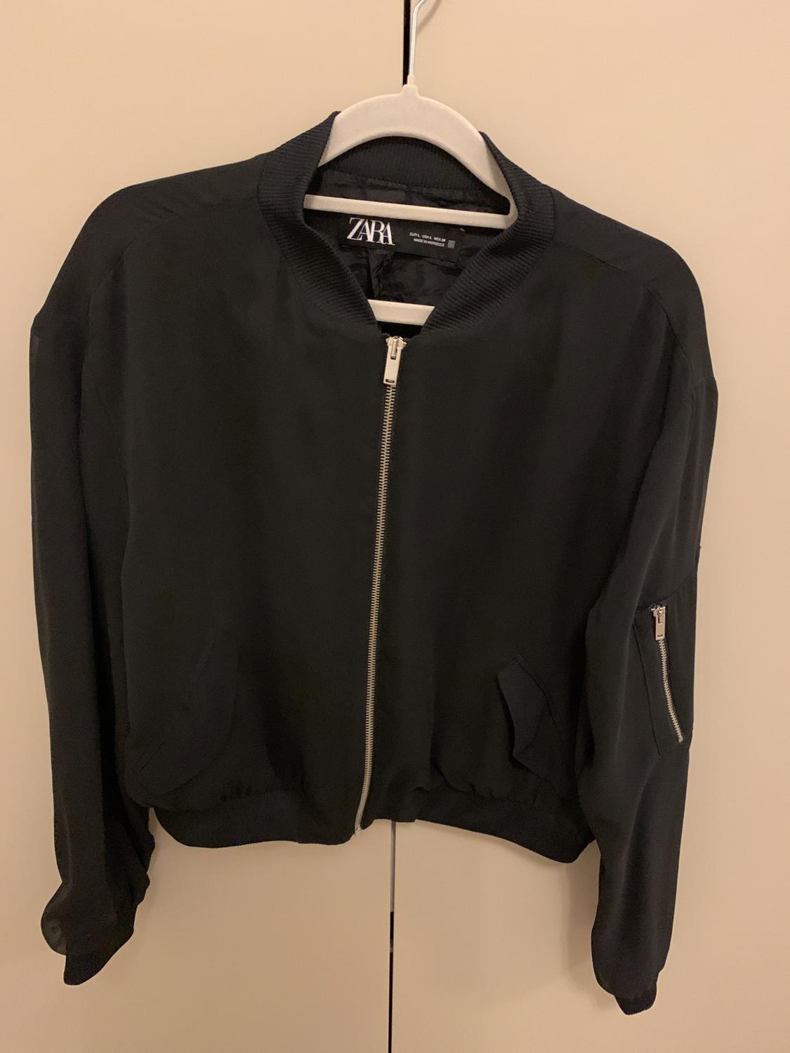 Black jacket with a zip and pockets, hanging on a white hanger. Visible label with the text "ZARA". Beige background.
