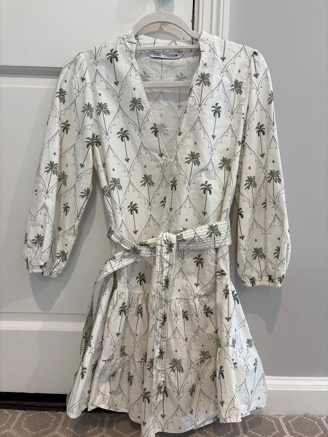 White short dress with green palm tree print, V-neck and belt, hanging on a hanger. Label with text ZARA.