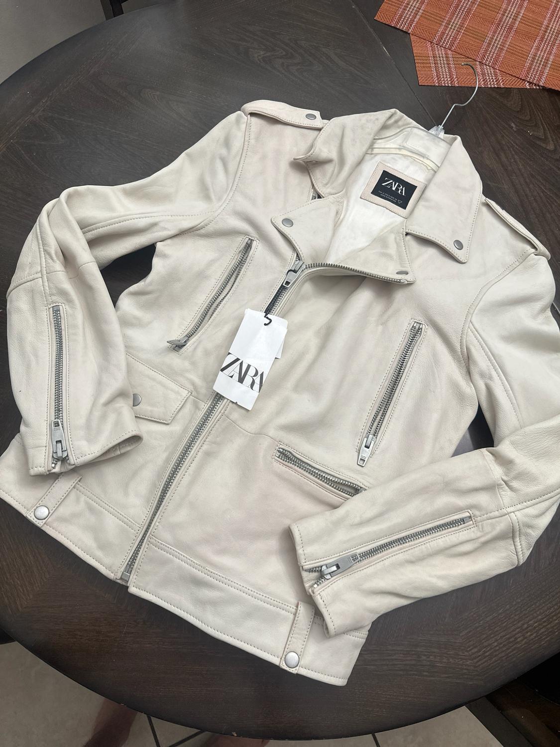 Cream-colored biker-style jacket with multiple silver zippers on a dark wooden table. Visible tag with the text ZARA.