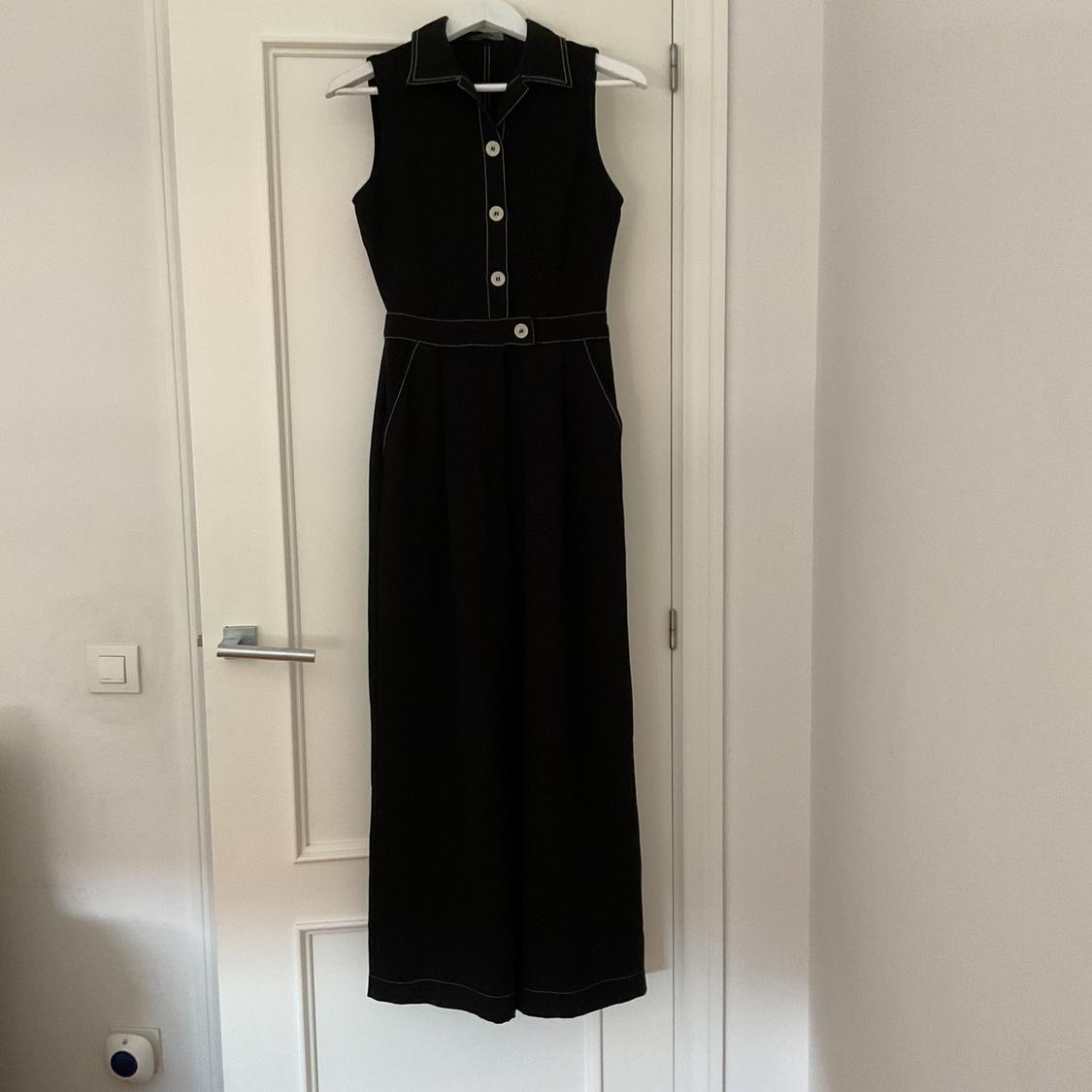 Sleeveless black jumpsuit with white buttons and visible stitching, hanging on a white door in an indoor setting.