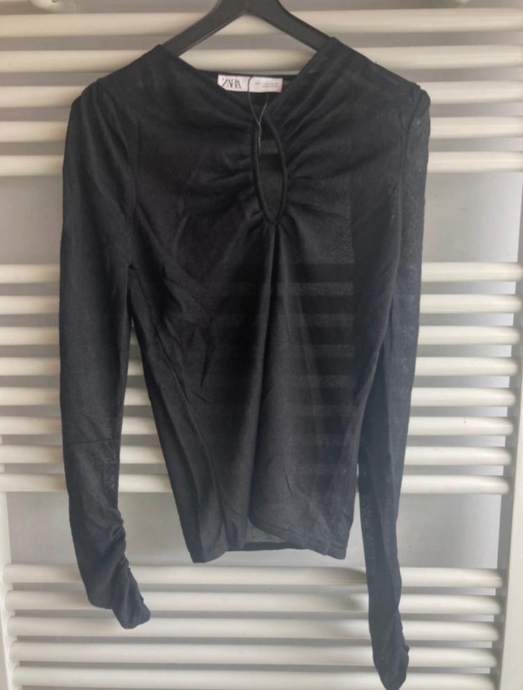 Black long-sleeved blouse hanging on a hanger, with a keyhole design at the neckline. Horizontal white slatted background.