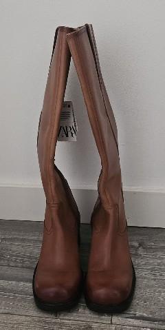 Brown high boots on a grey wooden floor in front of a white wall. They have a label with the text ZARA.