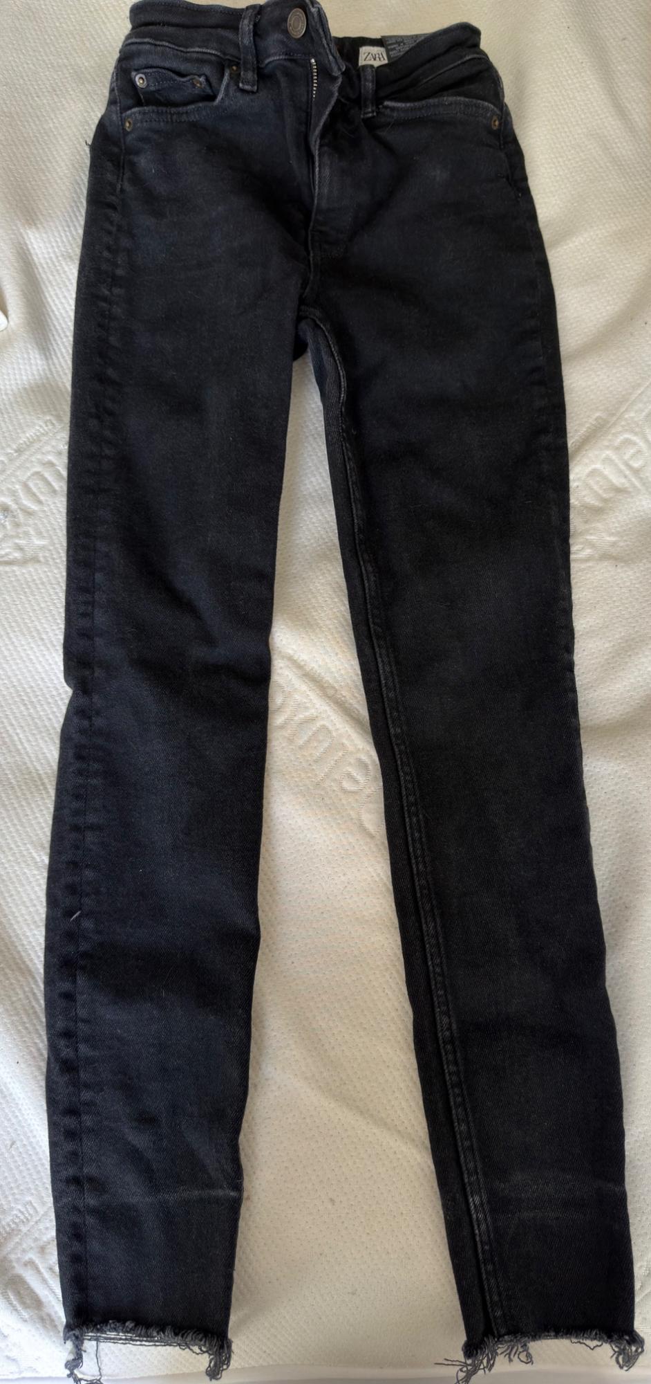 Black denim jeans with ZARA tag and frayed hem, laid flat on a textured white surface. Casual and relaxed style.
