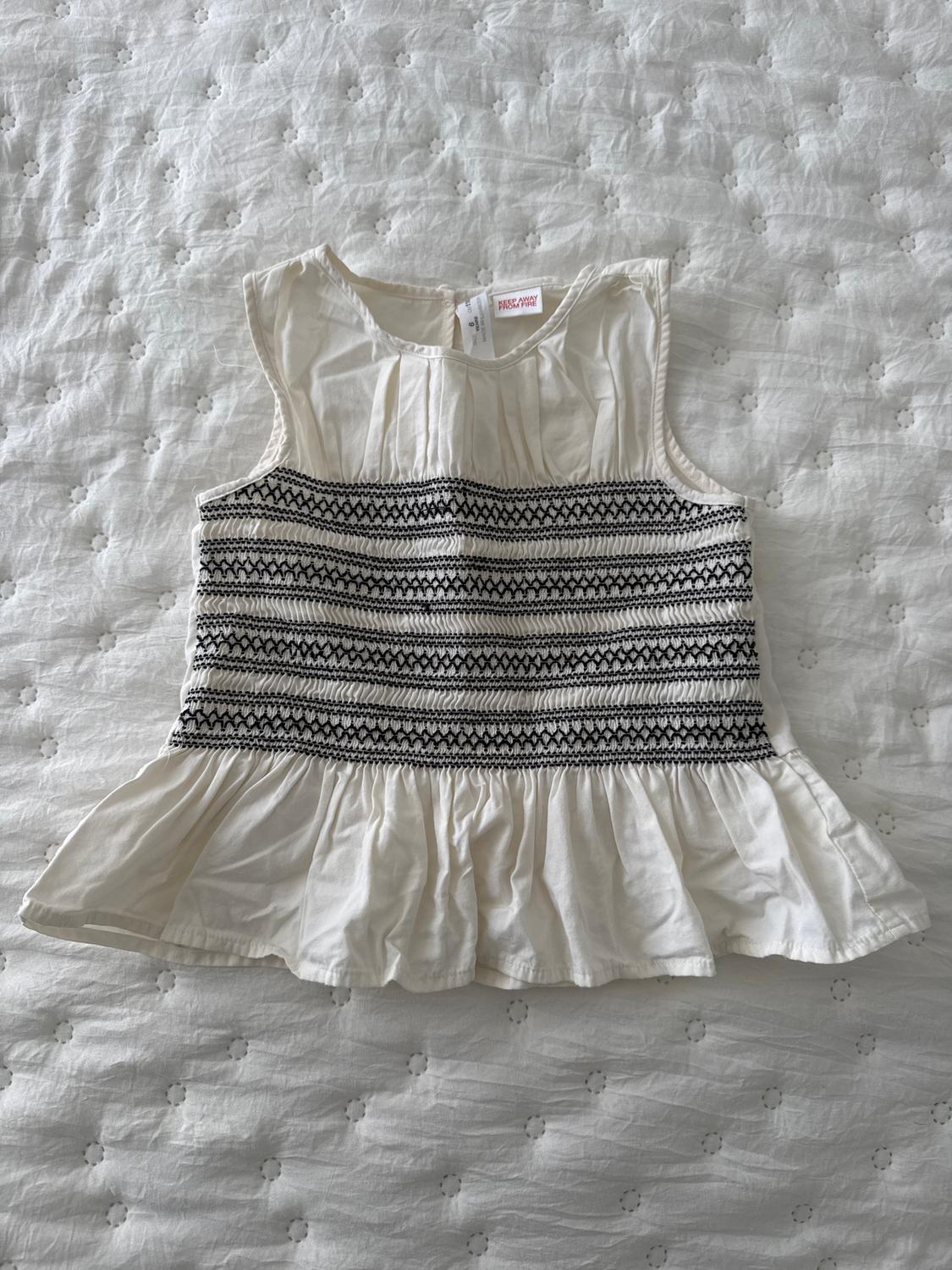 White sleeveless blouse with black embroidery on the chest and a frill on the hem, on top of a white quilted bedspread.