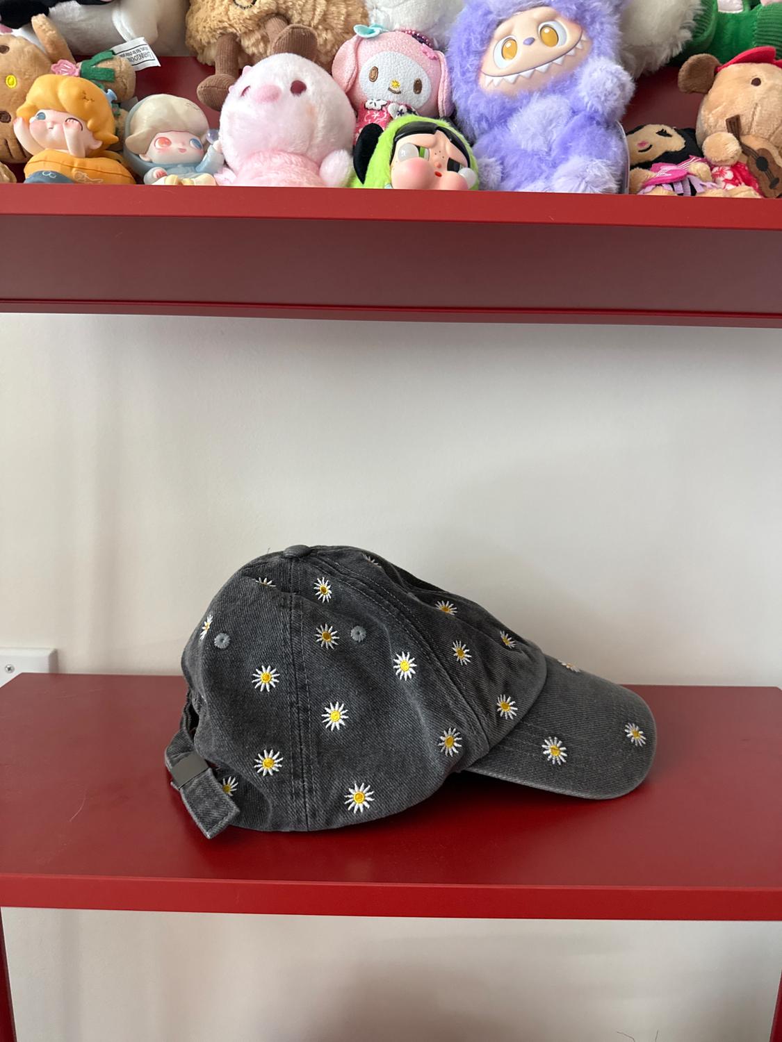 Dark gray cap with embroidered daisies on a red shelf. Above, a shelf with colorful stuffed animals. Casual and relaxed style.