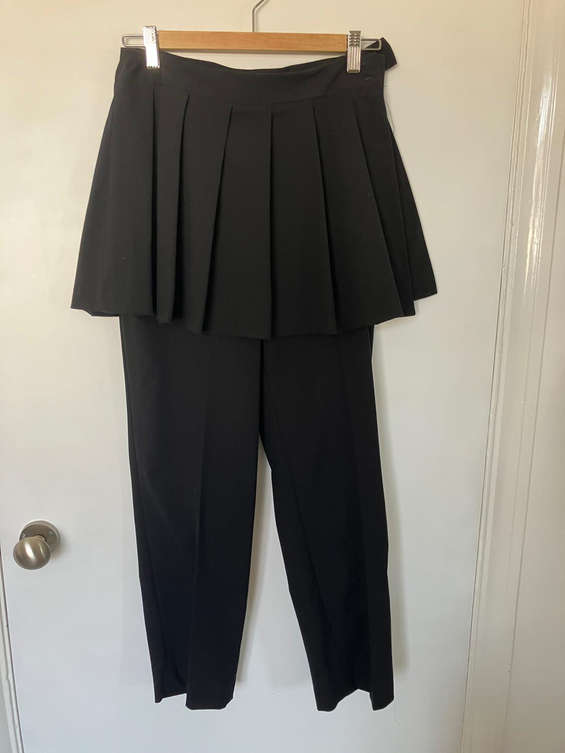 Black trousers with an overlaid pleated skirt, hanging on a wooden hanger over a white door.