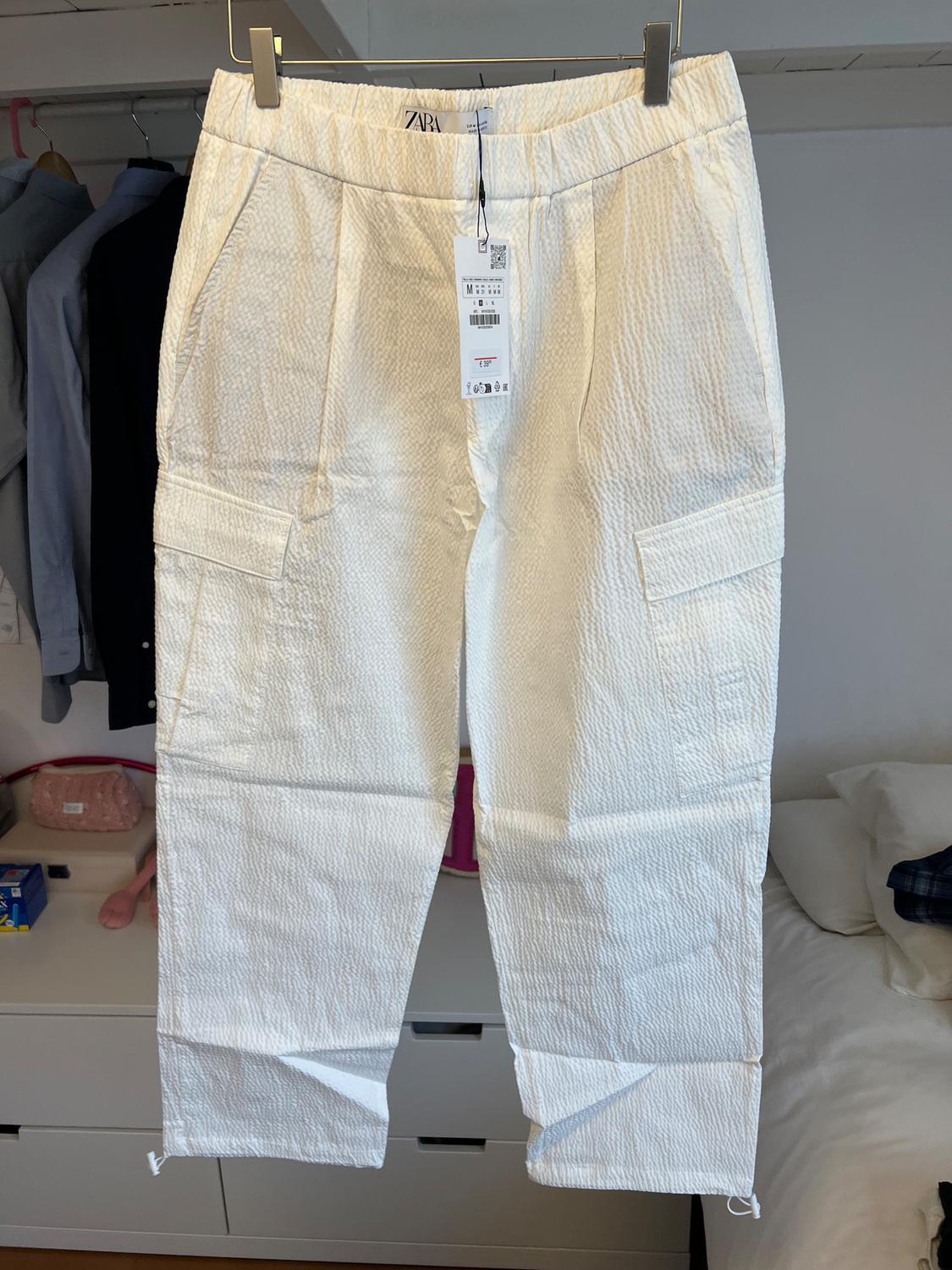 White textured fabric trousers hanging on a hanger, with a visible Zara tag, in a bedroom with clothes in the background.