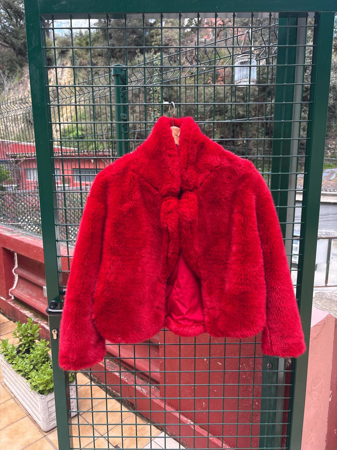 Red faux fur jacket hanging on a green metal door, with a garden and red building in the background.
