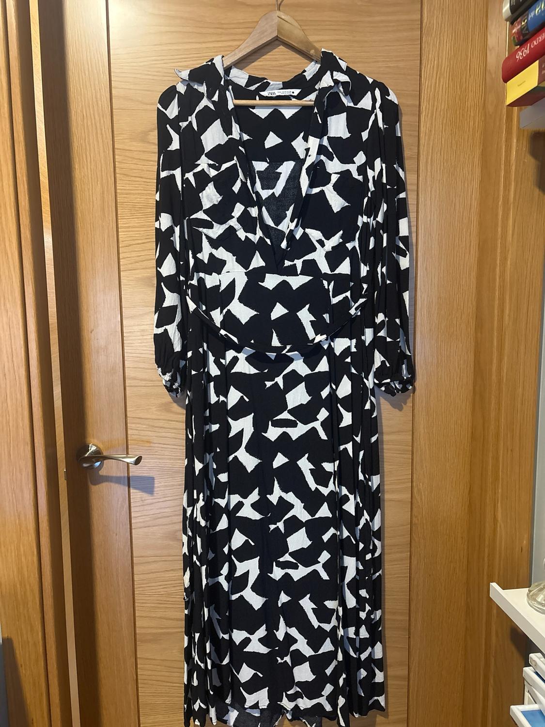 Black long-sleeved maxi dress with a white abstract print, hanging on a hanger over a wooden door. ZARA label.