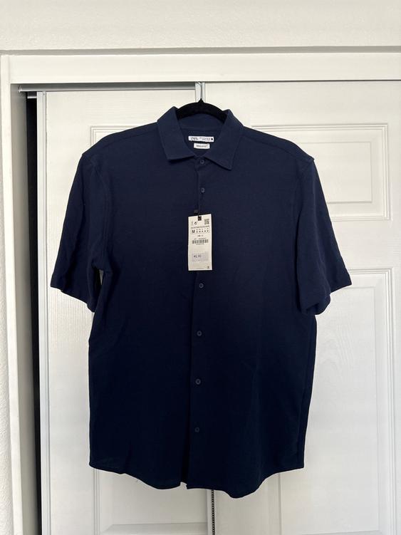 Navy blue short-sleeved shirt hanging on a hanger, with a visible price tag of 45.90, against a white door background.