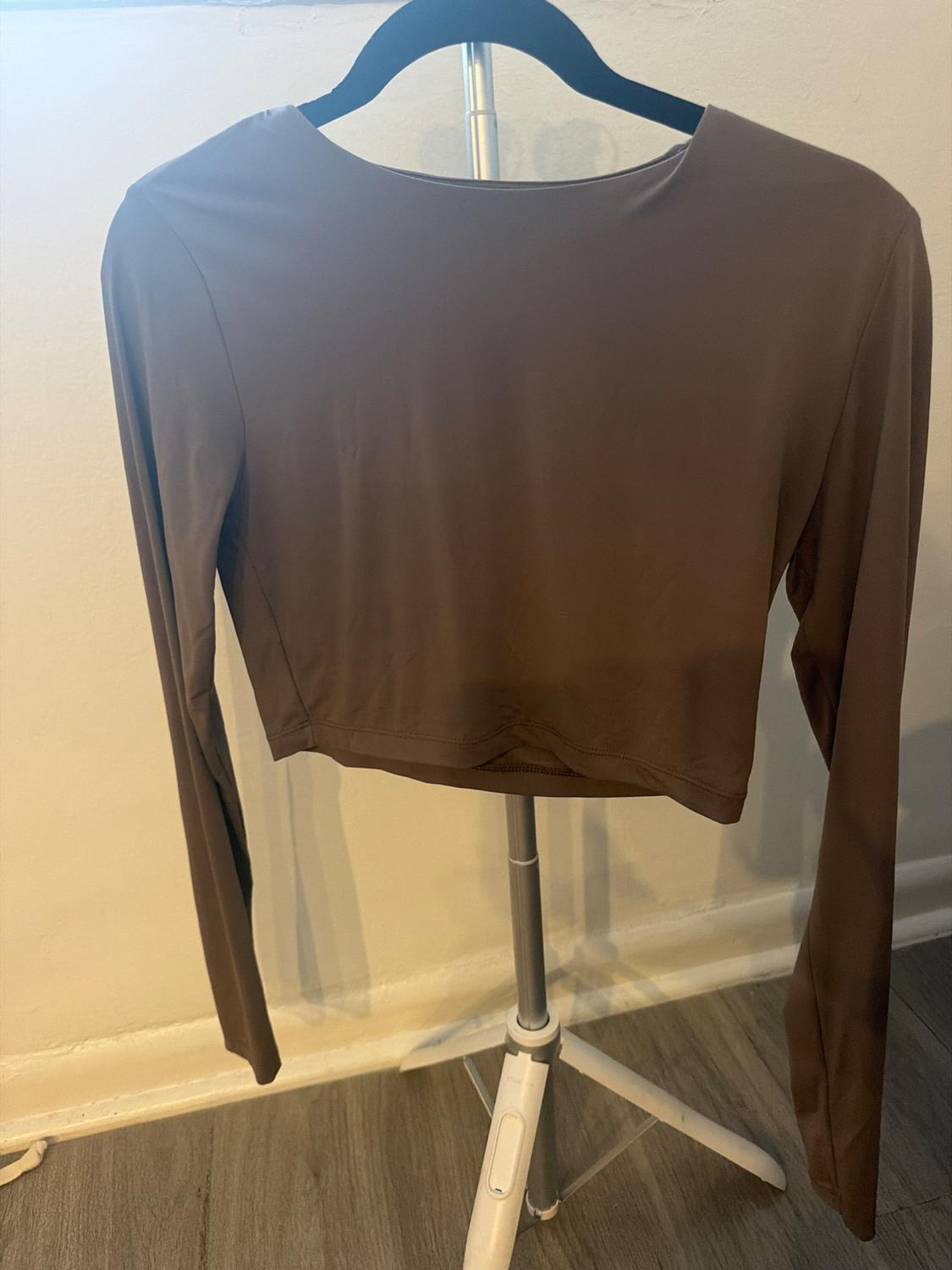 Brown long-sleeve crop top hanging on a black hanger on a tripod, in front of a white wall and a gray wood floor.
