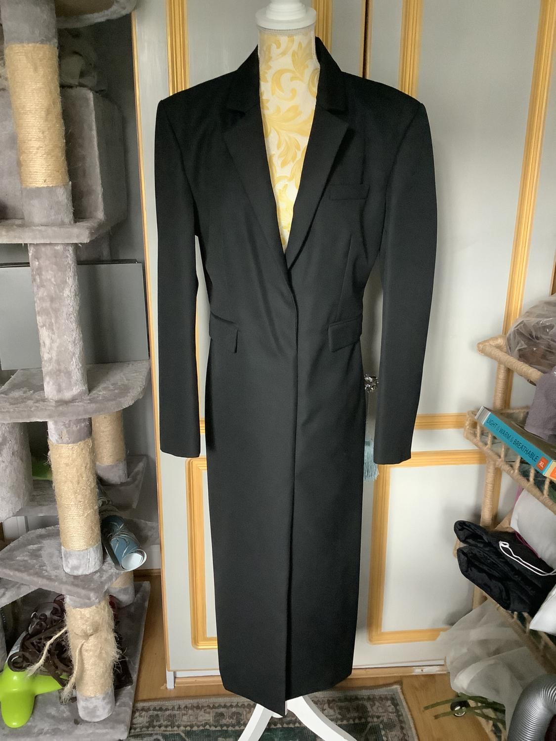 Long black coat on a mannequin with a bookshelf and cat scratcher in the background. Indoor setting with natural light.