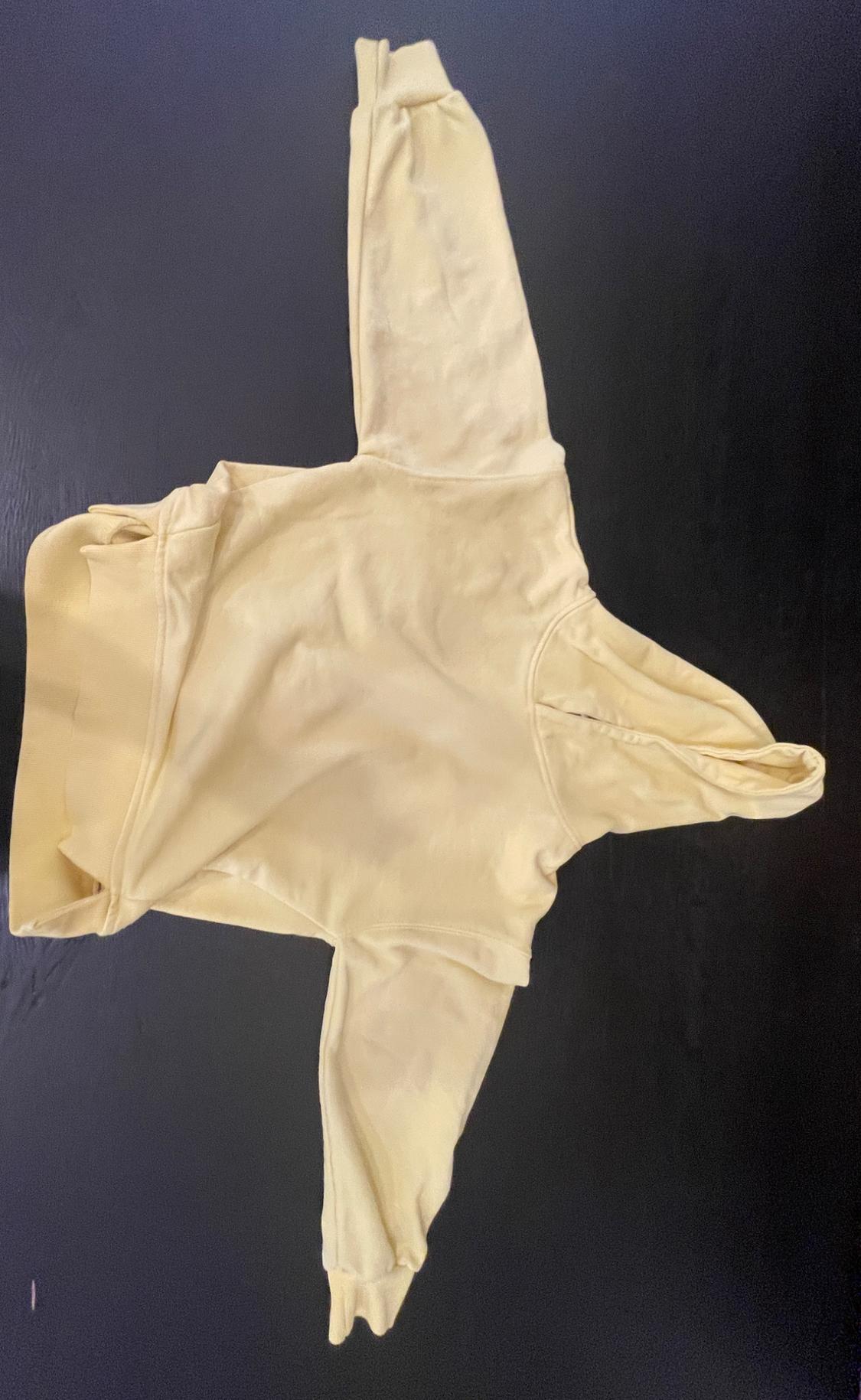 Light yellow hoodie laid out on a black surface. Casual and relaxed style with open sleeves.