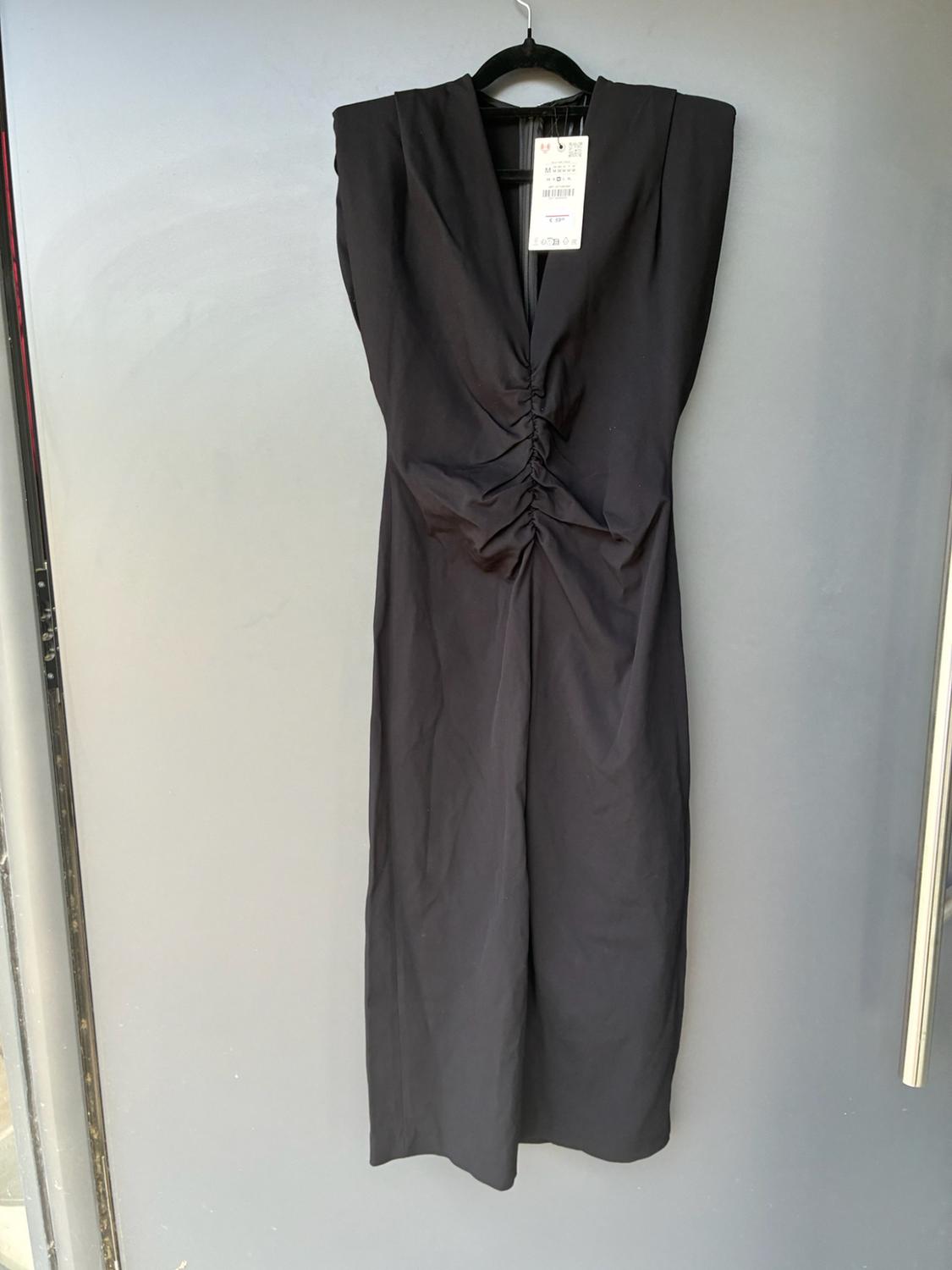 Sleeveless black dress with shoulder pads, a V-neckline and central ruched detail, hanging on a clothes hanger against a grey background.