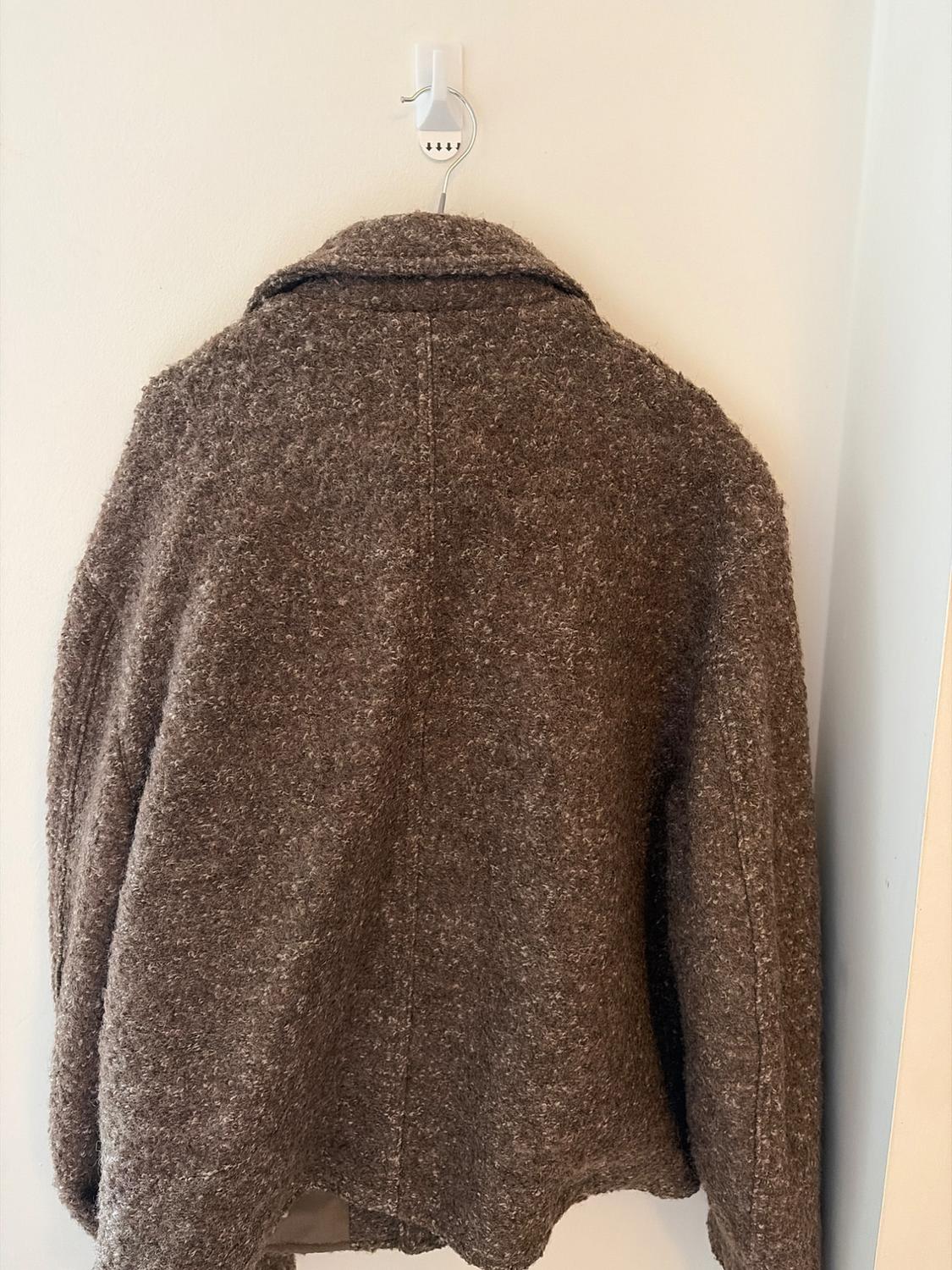 Thick-textured brown coat seen from the back, hanging on a hook on a white wall. Minimalist and relaxed atmosphere.