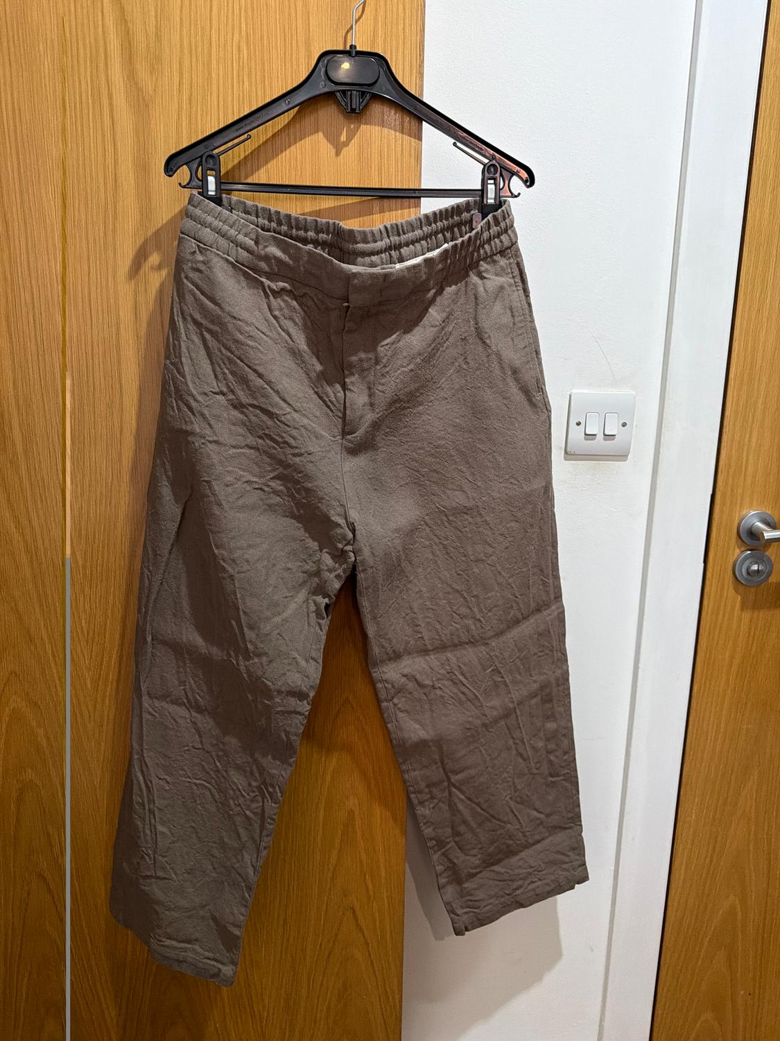 Wrinkled brown trousers hanging on a black hanger over a light wooden door, with a white wall in the background.