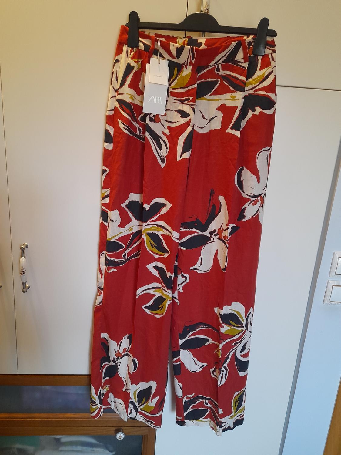 Red wide-leg trousers with a black and white floral print, hanging on a hanger in front of a white wardrobe. Label with the text ZARA.