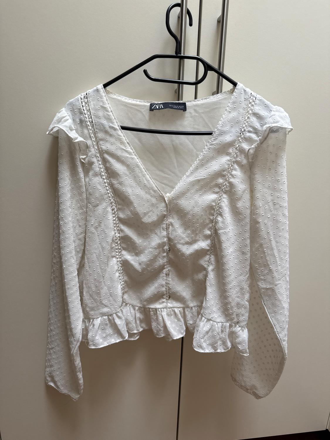 White blouse with a polka dot texture, long sleeves, and frill detailing on the hem, hanging on a black hanger in front of a wardrobe.