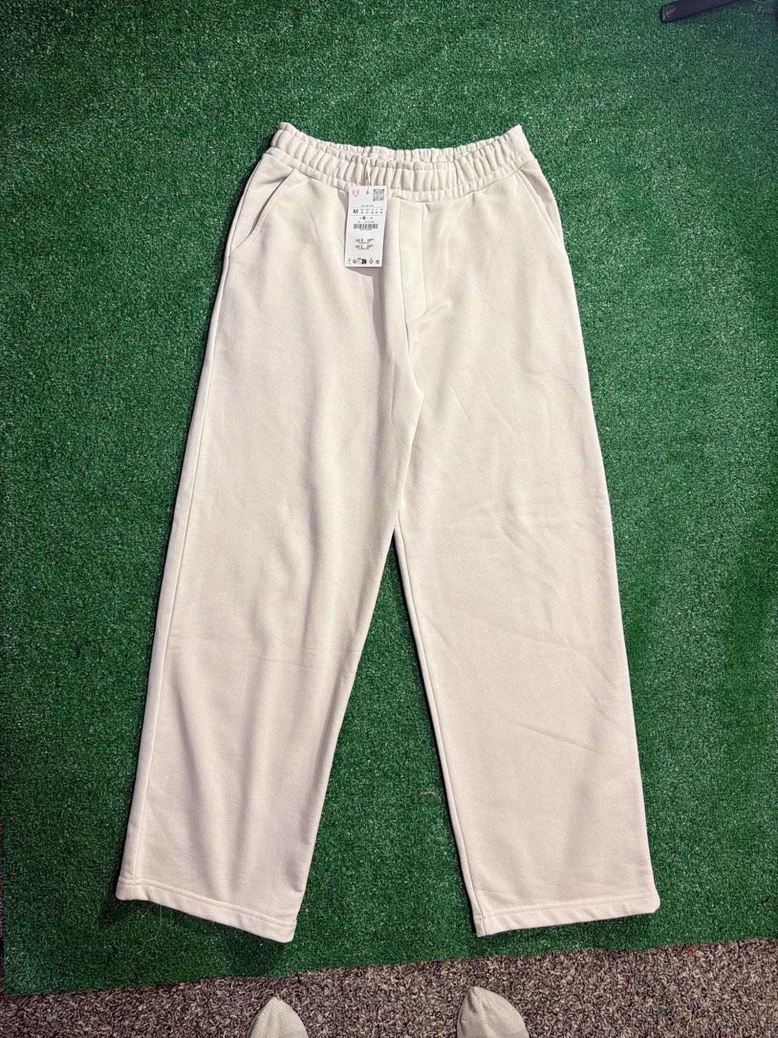 Beige wide-leg sweatpants with a tag, on green artificial grass. Feet wearing socks are visible along the bottom edge.