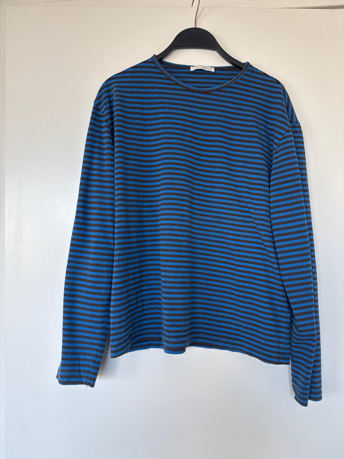 Blue and black horizontal striped long-sleeve t-shirt, hanging on a hanger with the text ZARA in front of a white background.