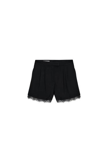 Front view of black Bermuda shorts with front pleats, side pockets and lace trim. Front zip fastening.