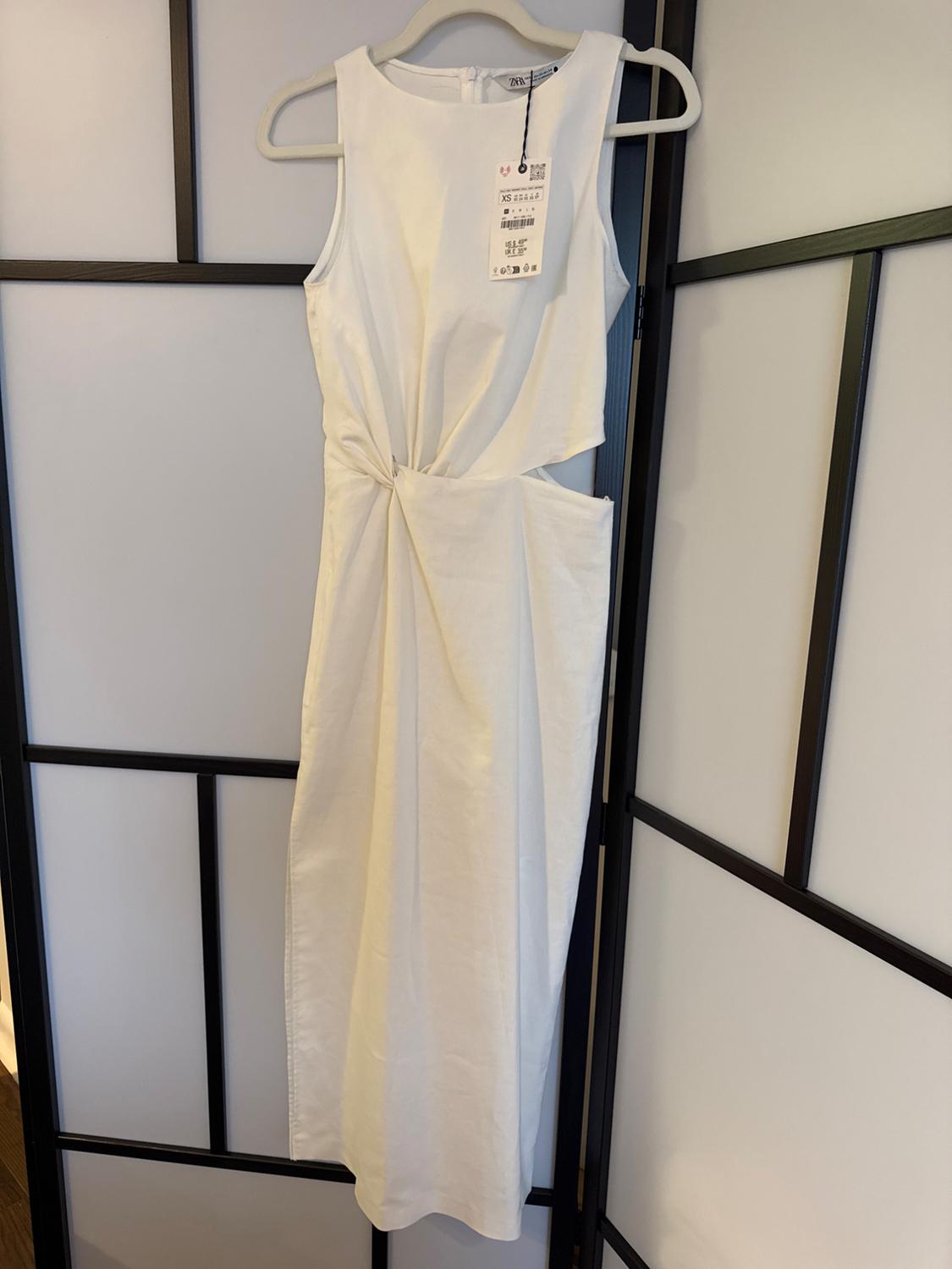 Sleeveless white dress hanging on a hanger, with a draped design at the waist. Visible tag with product details.