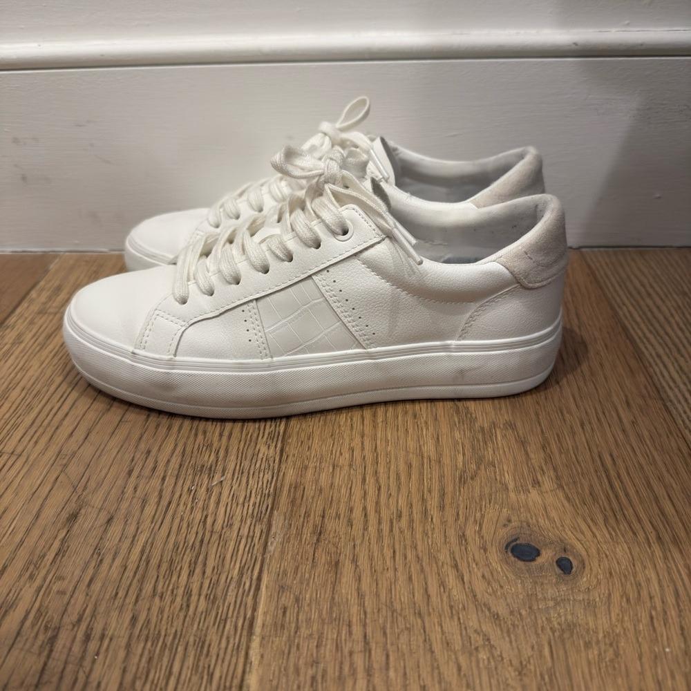 A pair of low-top white sneakers with laces and a textured side panel, placed on a wooden floor against a white wall.
